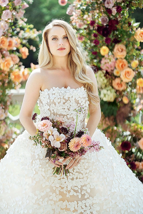Bridal portrait of a bride in a strapless floral lace gown holding a peach and blush bouquet with plum accents beneath a garden floral arch