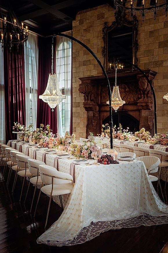 Reception tablescape with long banquet table wedding decor, pastel florals and taper candles beneath crystal chandeliers by a stone fireplace