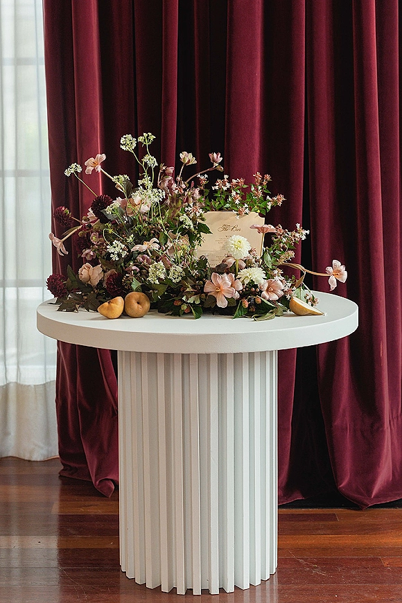 Wedding escort card display on a round pedestal table with escort cards, menu card, pears, and florals against burgundy velvet drapes
