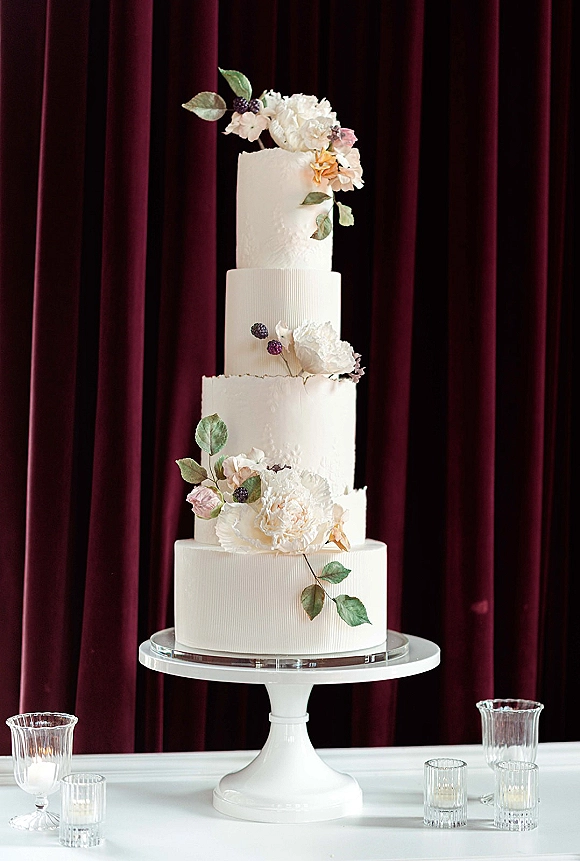 Wedding cake four tier wedding cake with textured white buttercream, sugar peonies, greenery and berries on a stand before burgundy velvet curtain