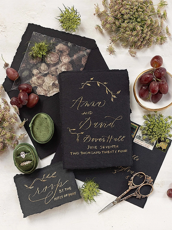 Wedding invitation suite with black deckle-edge paper and gold calligraphy, wax seal, rings, scissors, and grapes on white linen flatlay