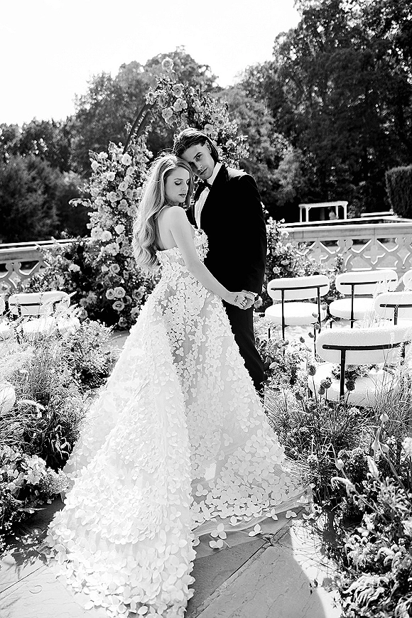 Couple portrait of bride and groom embrace, with a floral ceremony arch behind them on a garden terrace aisle with chairs and flowers