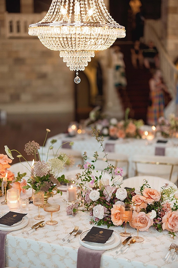 Reception tablescape with wedding table centerpiece of blush florals, taper candles and pink goblets beneath a crystal chandelier in stone venue