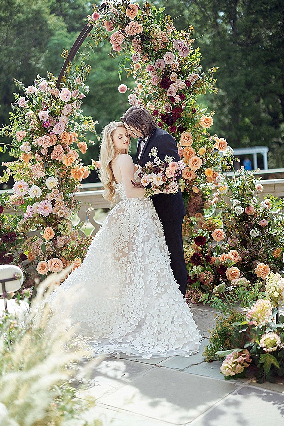 Couple portrait of bride and groom embrace beneath a lush floral arch, her rose bouquet and strapless gown on a garden stone patio