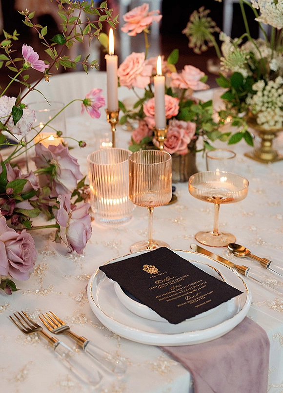 Reception tablescape with wedding place setting, pink and mauve roses in bud vases, brass candlesticks and taper candles among guests and chairs