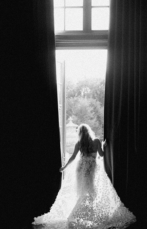 Bridal portrait of a bride from behind in a lace mermaid wedding dress with a long train, opening curtains at a bright window doorway.