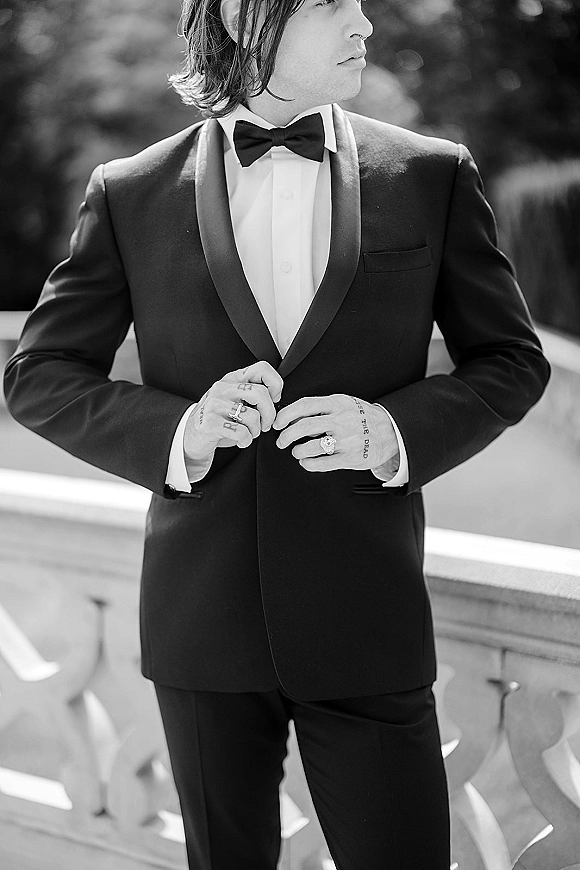 Groom portrait in a black tuxedo groom look, adjusting his jacket to show wedding band and tattoos by a stone balcony railing