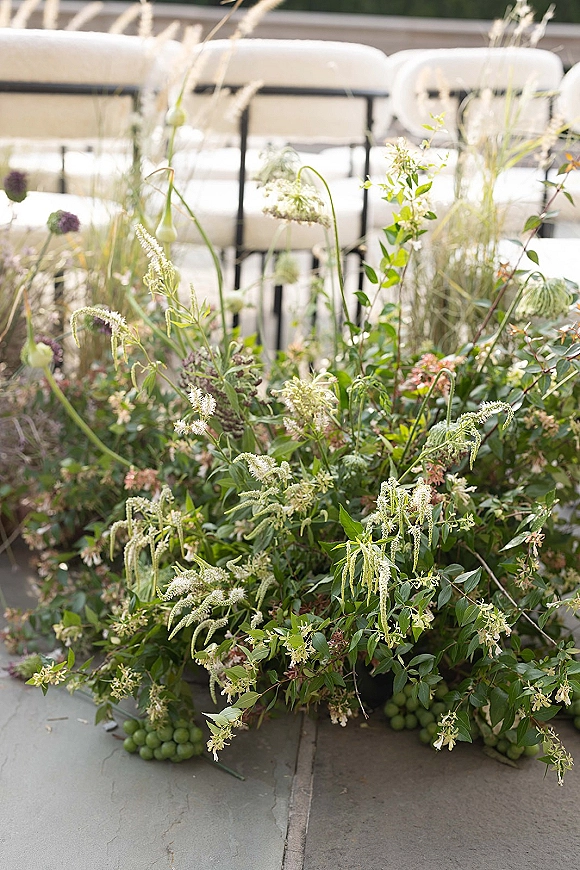 Ceremony aisle florals with meadow-style wildflower and greenery clusters with grape accents beside white chairs on a stone terrace
