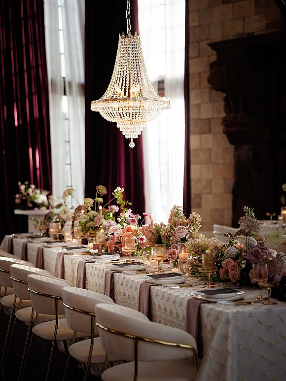 Reception tablescape with wedding banquet table set in mauve runner, low florals, votive candles, gold-rim glassware under crystal chandelier by fireplace