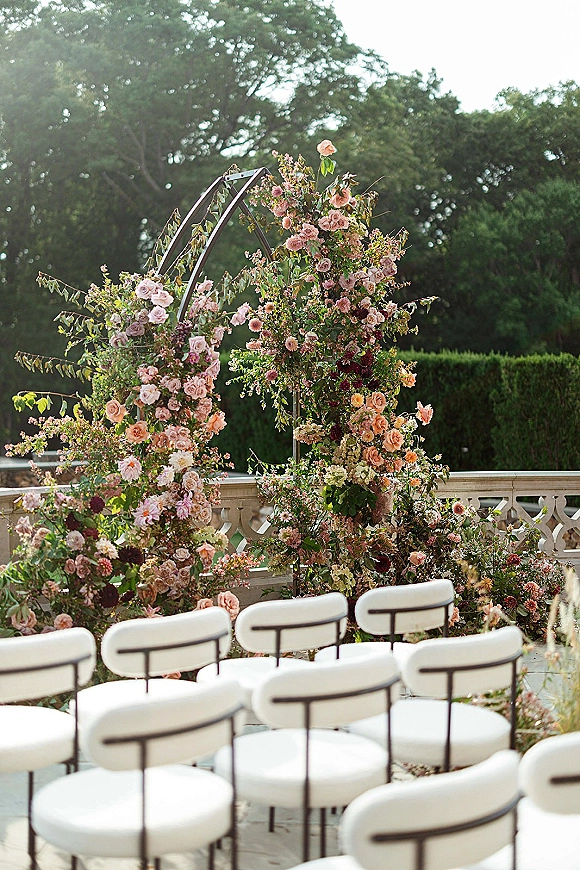 Ceremony arch decor with an asymmetrical floral arch of roses and climbing greenery on a metal frame, set on a garden terrace with white chairs