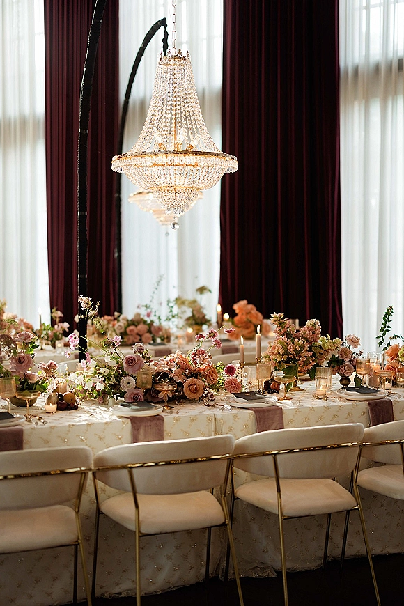 Reception tablescape with wedding head table decor, taper candles and floral centerpieces beneath a crystal chandelier by tall windows with burgundy drapes