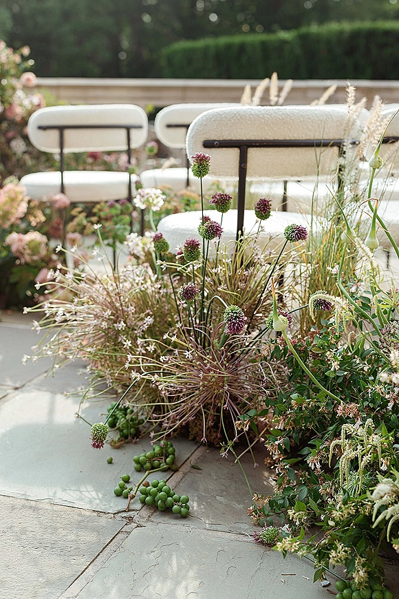 Ceremony aisle decor with outdoor ceremony chairs lined in white, flanked by meadow-style ground florals, allium and grasses on a stone patio