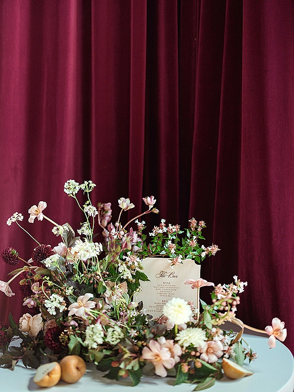 Bar menu sign with wedding bar menu card beside florals, greenery, pears and a lemon wedge on table against burgundy velvet curtain