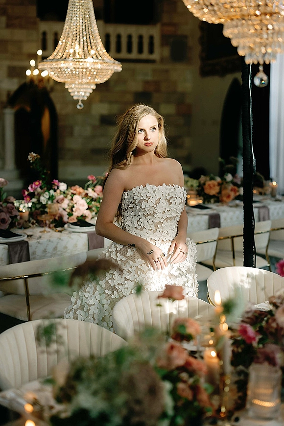 Bridal portrait of a bride in a strapless lace dress with floral appliqué, standing by candlelit reception tables beneath a crystal chandelier.