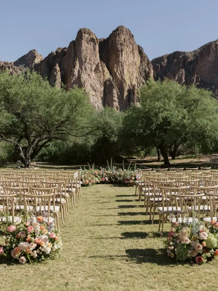 Ceremony setup for an outdoor wedding ceremony with wood chairs in a semicircle, wide aisle and pastel ground flowers on a desert lawn with rocky mountains
