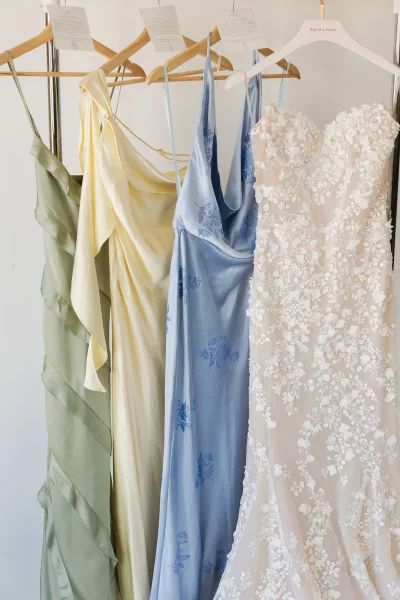 Wedding dress with lace appliqué hanging on a wooden hanger beside satin slip dresses and vow cards against a white wall