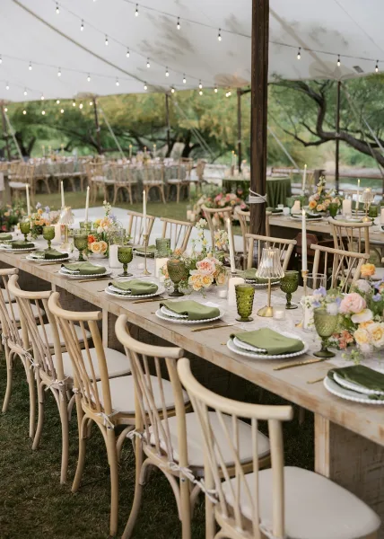 Reception tablescape for an outdoor wedding reception with farmhouse banquet tables, green goblets, gold flatware, candles, flowers under string lights by a river