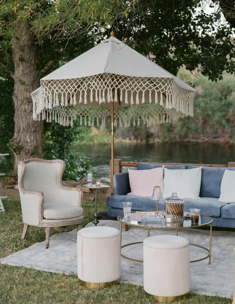 Outdoor lounge setup with wedding lounge furniture, featuring a blue velvet sofa under a fringe umbrella by a riverside lawn with trees