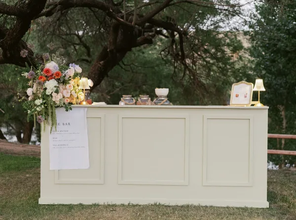 Wedding bar setup with floral arrangement, printed bar menu, and glass jar dispensers on a counter, lit by table lamps on a lawn by a wooden fence