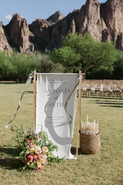 Wedding welcome sign on a wooden stand with ribbon streamers and floral arrangement, set by ceremony chairs on a grassy lawn with mountains beyond
