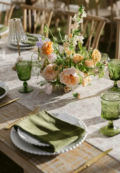 Reception tablescape with wedding table centerpiece of peach roses and white blooms, green goblets and napkins on a patterned runner outdoors