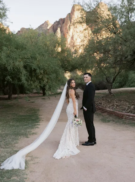 Couple portrait of bride and groom, her long veil trailing on the dirt path as they stand in a desert landscape with mountain rocks