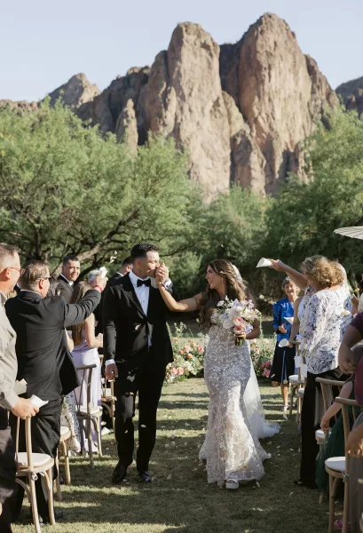 Wedding recessional as bride and groom walk the aisle holding hands, groom kissing her hand amid petals, with mountain landscape behind
