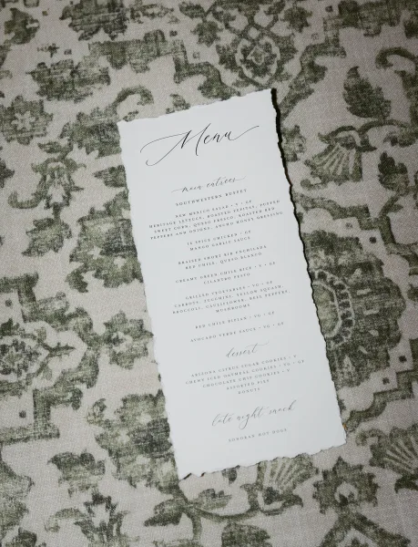 Wedding menu card with calligraphy on deckle edge paper, set on a patterned linen tablecloth with floral textile background