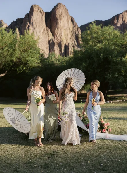 Bride with bridesmaids walking with bride across a grassy lawn, holding bouquets and white paper parasols under a blue sky by rocks