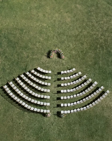 Ceremony seating layout with curved rows of white chairs and a floral arch with arrangements on a grass lawn, seen from above