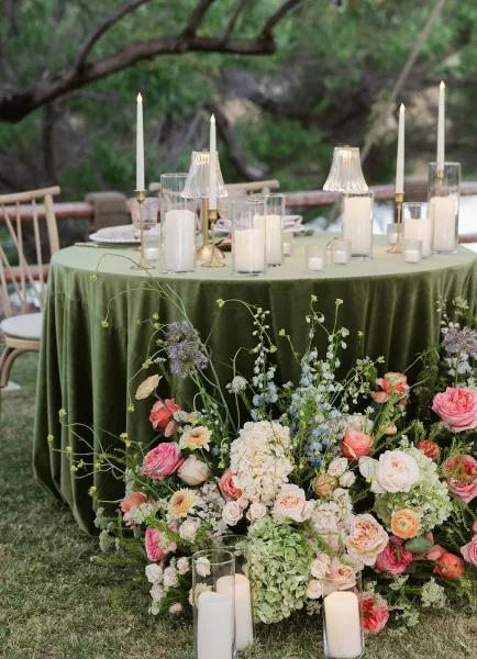 Reception tablescape with green velvet tablecloth, hydrangeas and roses, taper candles and glass hurricanes on a lawn by water and trees