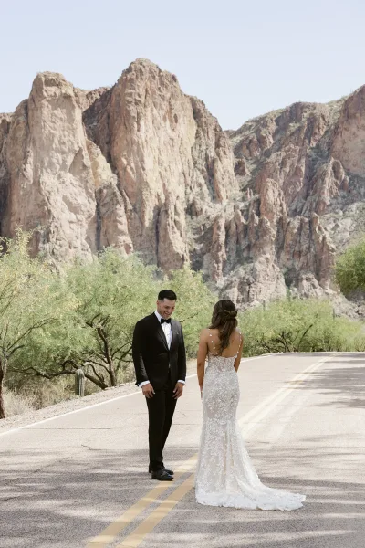First look moment as the bride in a lace gown with a long train walks up behind a tuxedoed groom on a desert road with mountains and cactus