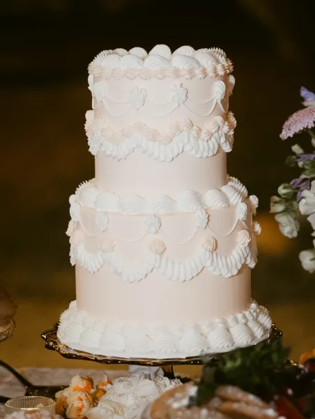 Wedding cake with scalloped buttercream piping, three-tier design on a cake stand with floral accents against a dark dessert table backdrop