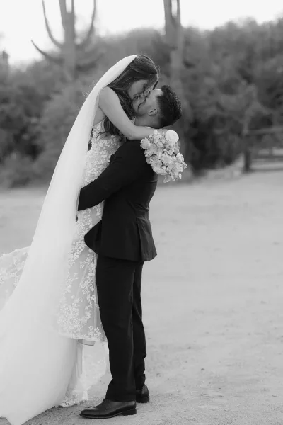 Wedding kiss portrait of groom lifting bride in a lace wedding dress with bridal veil on an outdoor tree-lined path