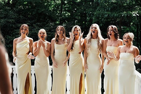Bridesmaid reveal as bridesmaids react in champagne satin dresses on an outdoor deck with white railing, surrounded by trees and greenery