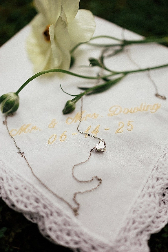 Wedding invitation suite with calligraphy wedding invitation and a heart pendant necklace accent, styled on lace with white flower buds and foliage
