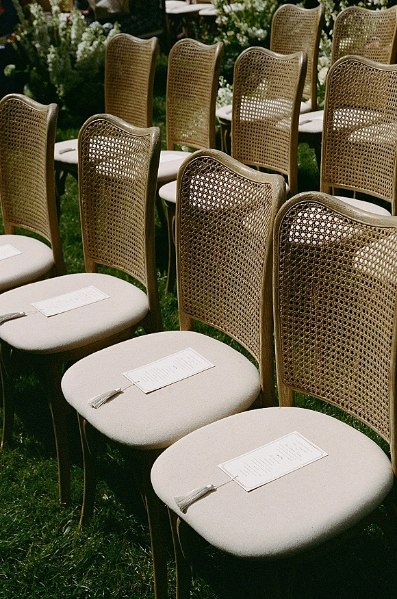 Ceremony seating with outdoor ceremony chairs featuring cane backs and cream cushions, programs with tassels, on a green lawn with florals