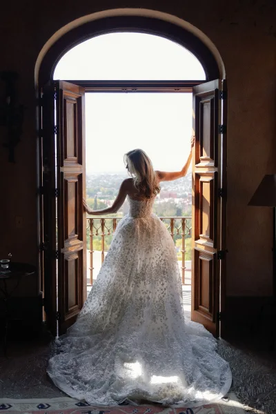 Bridal portrait of a bride from behind in a strapless lace wedding dress with a long train, standing by wooden doors to a balcony city view