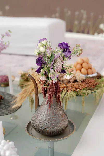 Wedding centerpiece with wildflower arrangement in a metal pitcher, purple blooms and dried amaranth on a dessert table with nuts and eggs
