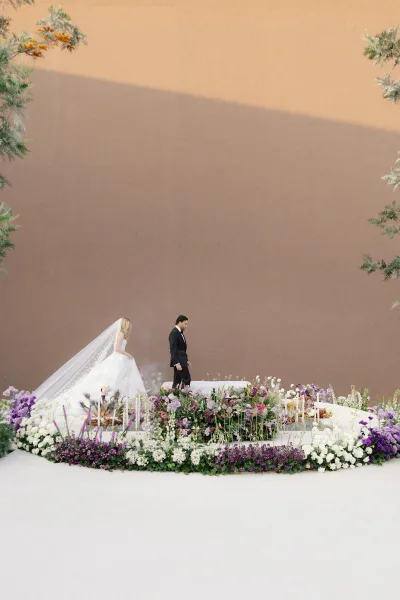 Ceremony moment as bride follows groom in tuxedo onto a white stage platform, cathedral veil trailing beside candlelit meadow florals.