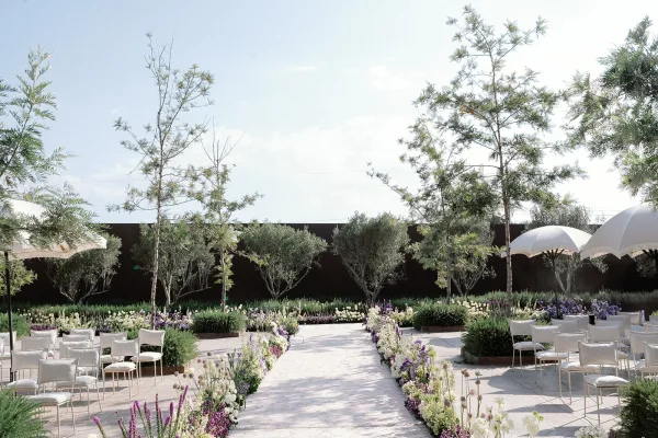 Ceremony aisle decor with a white aisle runner and purple and white ground florals, lined by white chairs and patio umbrellas in a garden courtyard