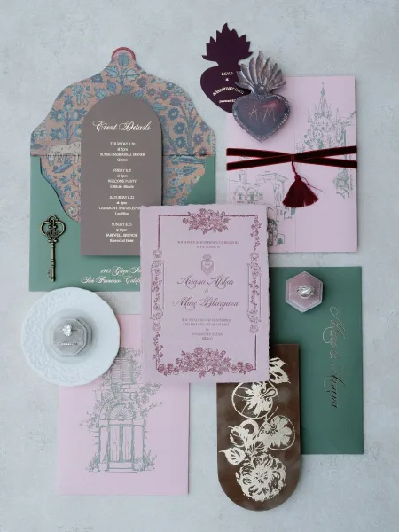 Invitation suite flat lay with pink wedding invitations, envelopes, RSVP and details cards, wax seal, velvet ribbon, and rings on gray surface