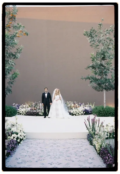 Wedding ceremony with bride and groom at a mountain altar, aisle runner bordered by purple-white florals and candles under sunset sky