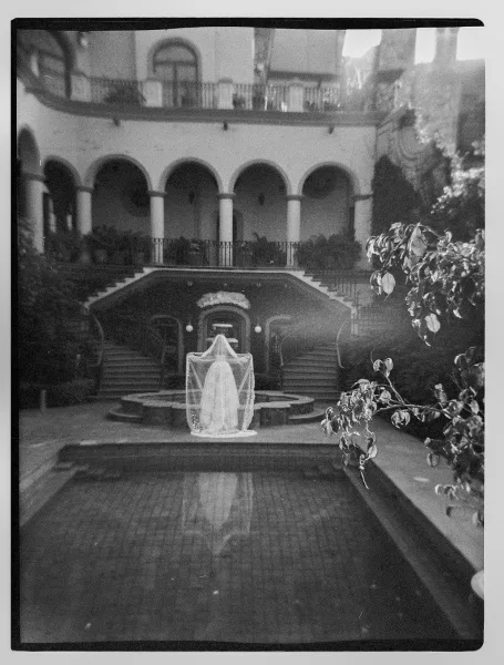 Bridal gown display with wedding dress hanging and veil draped in a courtyard by a reflecting pool, framed by stone arches and columns