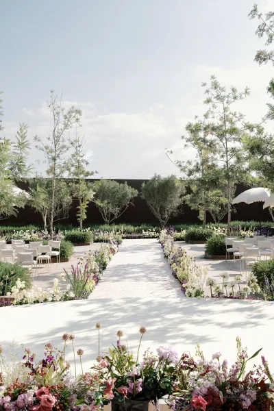 Ceremony setup with outdoor wedding ceremony aisle flowers lining a stone patio, white chairs and umbrellas framed by trees and hedges