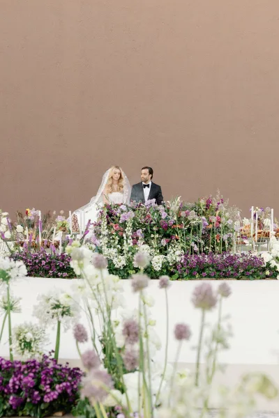 Sweetheart table decor with head table floral runner of wildflowers and greenery, white taper candles, as bride and groom sit by a neutral wall