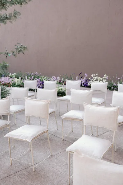 Ceremony seating with outdoor ceremony chairs in white, cushioned with linen, bordered by low florals against a stucco wall patio