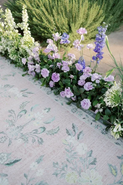 Ceremony aisle florals line a wedding aisle runner with lavender, blue delphinium, and white blooms on a stone garden walkway