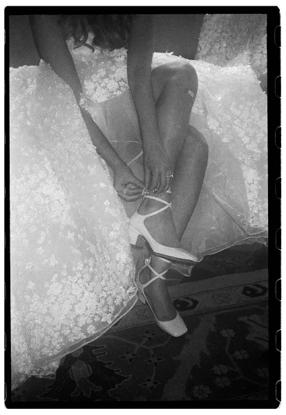 Bridal shoes with ankle strap heels as the bride ties ribbon straps beneath a lace wedding dress, seated on an armchair over a patterned rug