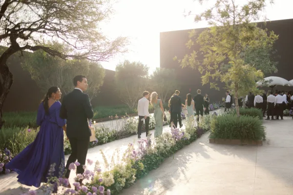 Wedding cocktail hour with wedding guests arriving in cocktail attire, mingling along a flower-lined garden walkway on a sunny patio